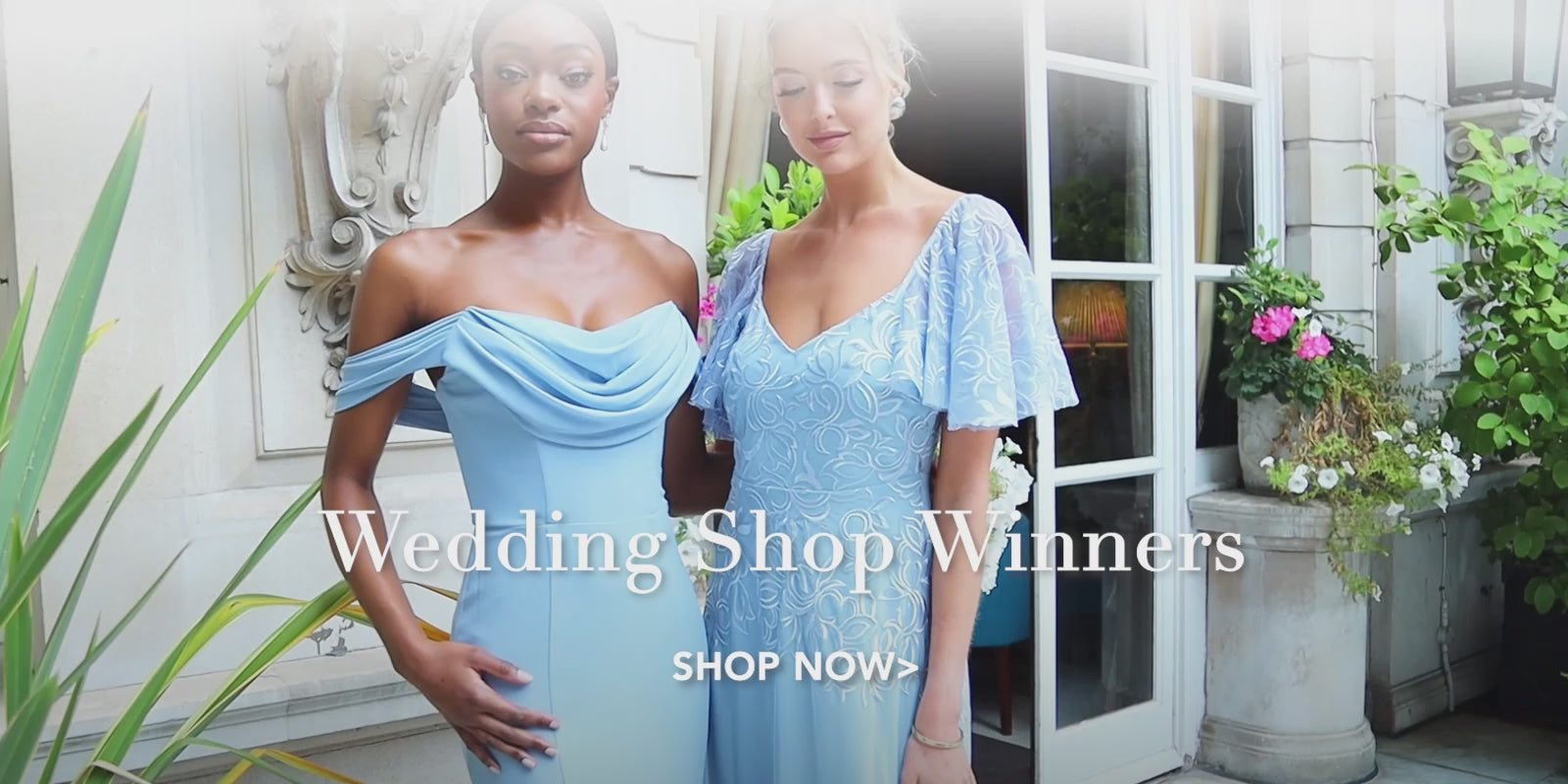 Two bridesmaids wearing elegant light blue maxi dresses pose gracefully in front of a charming wedding backdrop featuring a house adorned with flowers and lush green plants.